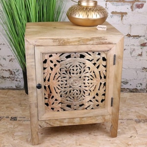 May include: A light-coloured wooden bedside table featuring a carved floral design on the door. The table has a small black knob and is topped with a gold-coloured decorative vase. The table is placed against a brick wall.