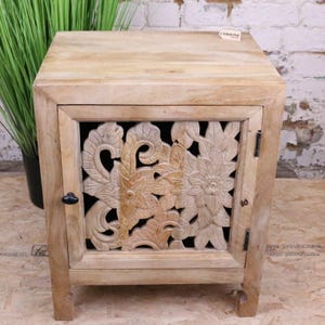 Puede incluir: Un mueble de madera clara con un diseño floral tallado en la puerta. El mueble tiene una sola puerta con un pomo negro y está sobre cuatro patas cortas. La madera tiene un acabado natural.