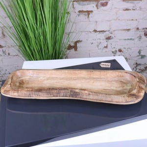 May include: A rectangular, light brown wooden bowl with a carved interior. The bowl has a natural wood grain pattern and is set against a white and grey background. The bowl is approximately 50 cm long.