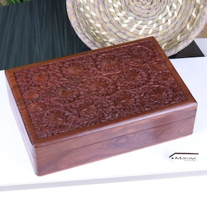 May include: A rectangular wooden box with intricate floral carvings on the lid. The box is made of dark brown wood and has a polished finish.
