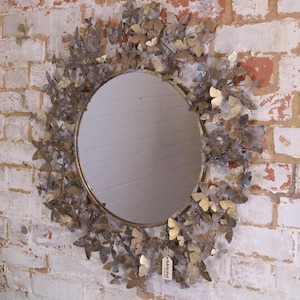 May include: A round mirror with a gold-colored frame, surrounded by numerous metal butterflies in shades of gold and silver. The mirror is mounted on a white brick wall, creating a decorative accent.