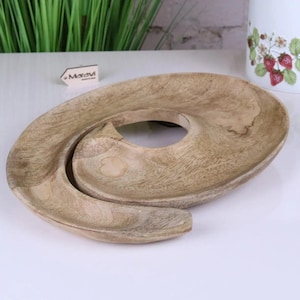May include: A light brown, wooden serving tray with a unique, spiral design. The tray features a central circular opening and a curved, elongated shape. The wood grain is visible, adding texture. The tray is suitable for serving snacks or decorative items.