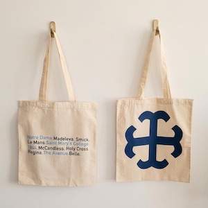 May include: Two off-white canvas tote bags with two handles each. The left bag features black and blue text reading: Notre Dame. Madeleva. Smick. Le Mans. Saint Mary's College. 1844. McCandless. Holy Cross. Regina. The Avenue. Belle. The right bag has a large blue cross.