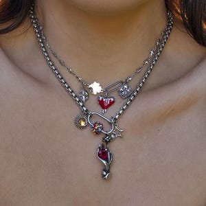 May include: A layered silver necklace with multiple charms. Charms include a red heart, a star, a sun, and a hand holding a red gem. The necklace is worn over a white top.