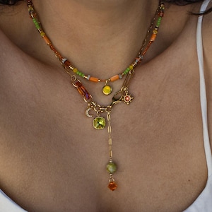 Può includere: Una collana a strati con perline colorate e ciondoli dorati. La collana presenta perline arancioni, verdi e gialle, con un pendente a forma di gemma quadrata verde e un piccolo ciondolo a forma di sole. La collana è indossata su una canotta bianca.