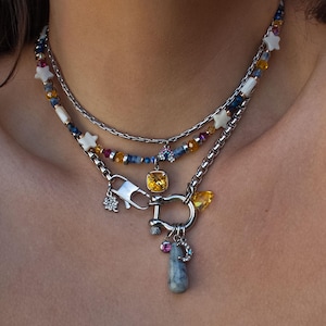 Peut inclure: Ensemble de colliers superposés avec des chaînes argentées et des rangées de perles colorées et de breloques en forme d'étoiles. La couche inférieure comprend un grand fermoir avec un pendentif en pierre précieuse et d'autres breloques décoratives. Le collier est porté sur un haut blanc.