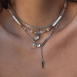 Könnte beinhalten: Ein mehrlagiges Halsketten-Set mit mehreren Strängen. Der obere Strang hat kleine Perlen und weiße Akzente. Der mittlere Strang hat einen quadratischen Anhänger. Der untere Strang hat eine Kette mit einem hängenden Stachel-Anhänger. Die Halsketten sind silberfarben.