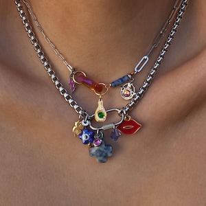 May include: Two layered silver necklaces with various charms. The top necklace has a chain link design, while the bottom necklace has a classic chain. Charms include a red lip, a cross, a four-leaf clover, and a small bottle.