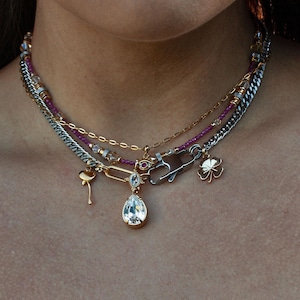 Könnte beinhalten: Mehrreihige Halskette mit goldenen, silbernen und Perlenketten. Die Halskette hat Anhänger, darunter ein tropfenförmiger Anhänger, ein Flamingo, ein vierblättriges Kleeblatt und einen Sicherheitsnadelverschluss. Die Halskette wird getragen.