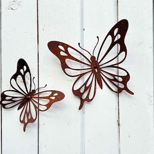 Può includere: Due decorazioni da parete a farfalla in metallo. Le farfalle sono in metallo marrone arrugginito e sono montate su uno sfondo di legno bianco. Le farfalle hanno intricati disegni ritagliati e sono di diverse dimensioni.