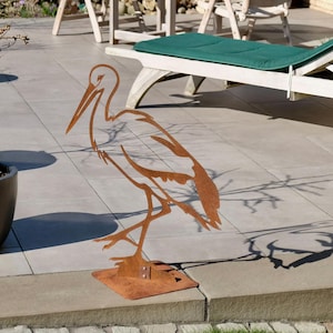 Op de afbeelding: Een roestig metalen beeld van een ooievaar die op een voet staat. Het beeld staat buiten op een terras.