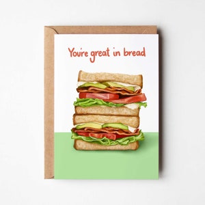May include: A greeting card featuring a vibrant illustration of a layered sandwich. The red text on the card reads "You're great in bread". The sandwich is made with lettuce, tomato, avocado, and bacon between toasted bread. The card is presented in a brown envelope.
