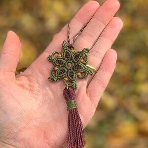 Könnte beinhalten: Ein grüner und burgunderfarbener Makramee-Schneeflocken-Anhänger mit einem burgunderfarbenen Quasten. Die Schneeflocke ist mit kleinen Perlen besetzt und hat ein filigranes, kompliziertes Design.