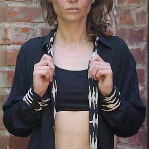 May include: Black button-down shirt with white and beige patterned accents on the cuffs and lapel. The shirt is worn open over a black crop top and dark denim jeans. The background is a brick wall.