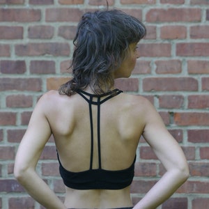 Può includere: Reggiseno sportivo nero con un esclusivo design a spalline sulla schiena. Il reggiseno presenta uno stile racerback con più spalline sottili che convergono al centro. La modella è vista da dietro, con un muro di mattoni sullo sfondo.
