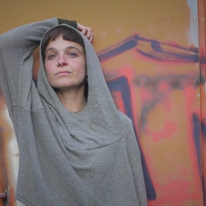 Könnte beinhalten: Eine Person trägt ein graues Kapuzenpullover mit der Kapuze hochgezogen. Die Person steht vor einer Wand mit Graffiti.