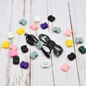 以下が含まれることがあります： カラフルなケーブルオーガナイザーと充電ケーブルの詰め合わせ。オーガナイザーは、ピンク、黄色、緑、紫、黒、白など、さまざまな色があります。ケーブルは黒色で、USB-AおよびUSB-Cコネクタが付いています。「iPhone Alice」というテキストが入った白い正方形もあります。