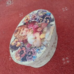 May include: Oval wooden box with a decoupage design featuring a young girl holding flowers. The flowers are pink, purple, and yellow. The background is white with a distressed look.