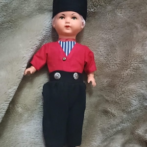 May include: A vintage doll dressed in traditional Dutch clothing. The doll has a black hat, red shirt, blue and white striped undershirt, black trousers, and wooden shoes.