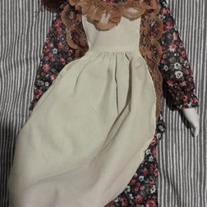 Peut inclure: Poupée en porcelaine avec des cheveux bruns et une robe à imprimé floral. La poupée porte un tablier beige avec une bordure en dentelle marron et des bottes marron. Le visage de la poupée a des traits peints, dont les yeux fermés et les lèvres rouges.