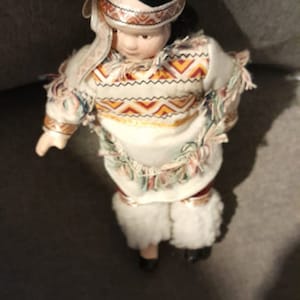 Peut inclure: Poupée en porcelaine vêtue d'une tenue amérindienne. La poupée a des cheveux noirs, un bandeau et une tenue crème avec des motifs géométriques colorés. La tenue comprend un haut à franges et des jambières en fausse fourrure blanche.