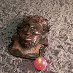 May include: A dark brown wooden mask with a carved human face. The mask has a geometric design and is sitting on a grey fluffy surface. A pink ball with gold glitter is in the foreground.