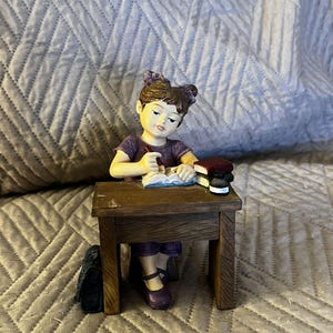 May include: A figurine of a girl sitting at a wooden desk, wearing a purple shirt and holding a book. She has brown hair with purple bows and is wearing purple shoes. There are two books stacked on the desk.
