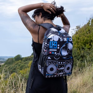 Peut inclure: Un sac à dos noir avec un design radio des années 80 coloré. Le sac à dos présente une cassette bleue et blanche, un tourne-disque et le texte "80's Radio Countdown".
