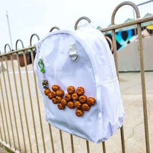 Op de afbeelding: Witte rugzak met een mesh-vak en een ritssluiting. De rugzak heeft een decoratief ontwerp van kleine, ronde, oranje-bruine objecten.