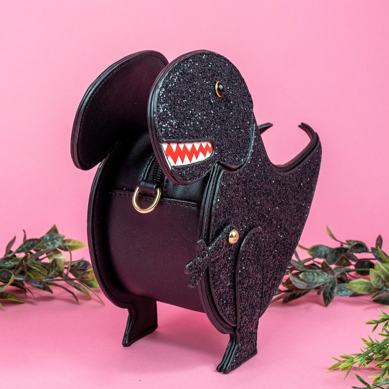 The kawaii black glitter dino vegan bag on a pink studio background with green foliage surrounding it. The bag is angled left and forward to highlight the dinosaur face, moveable arm, detachable strap and black glitter front.