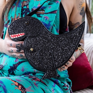 The kawaii black glitter dino vegan bag being held up by a tattooed model wearing dino print dungarees. The bag is facing forward to highlight the dinosaur face, moveable arm, detachable strap and black glitter front.