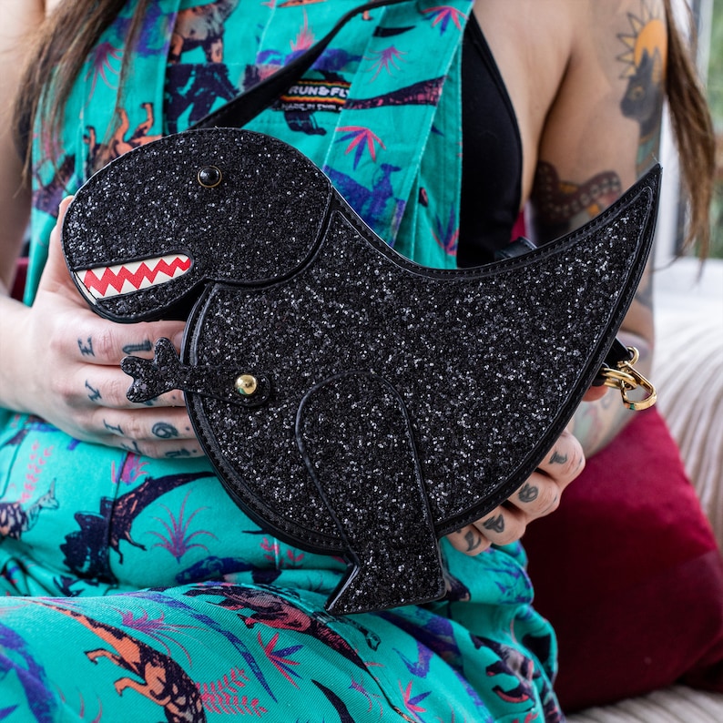 The kawaii black glitter dino vegan bag being held up by a tattooed model wearing dino print dungarees. The bag is facing forward to highlight the dinosaur face, moveable arm, detachable strap and black glitter front.