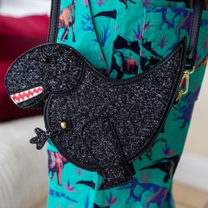 The kawaii black glitter dino vegan bag being worn by a model wearing dino print dungarees. The bag is facing forward to highlight the dinosaur face, moveable arm, detachable strap and black glitter front.