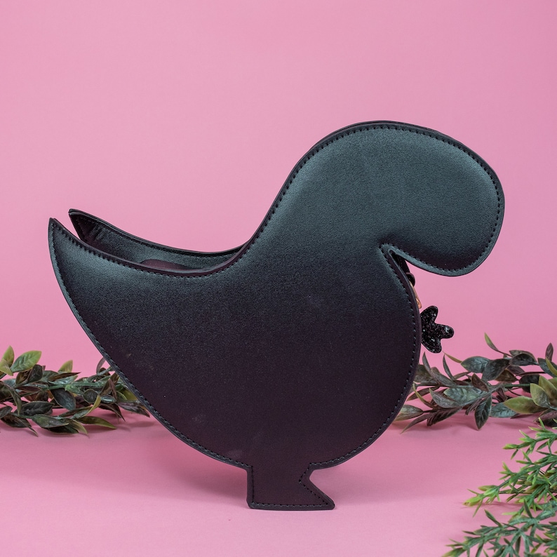 The kawaii black glitter dino vegan bag on a pink studio background with green foliage surrounding it. The bag is facing away from the camera to highlight the plain black back and detachable strap.