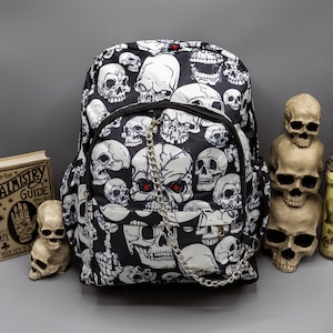 May include: A black backpack featuring an all-over white skull print. The bag includes a front zippered pocket, a silver-toned metal chain attached to the front, and a central skull graphic with two small red dots positioned as eyes on the front panel.