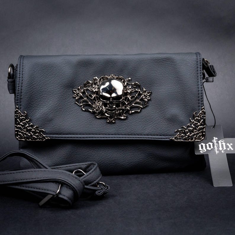 Gothic black vegan leather bag by GOTHX with a silver skull and rose motif, styled on a dark black background. Showing the cross tag and the detachable adjustable strap.