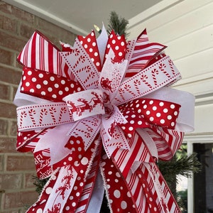 May include: A red and white Christmas tree topper bow with a variety of patterns including polka dots, stripes, and candy canes. The bow is tied with a white ribbon and has a frayed edge.