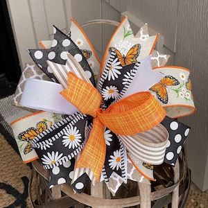 May include: A black and white polka dot, daisy, and butterfly patterned bow with orange and brown ribbon. The bow is tied around a wooden basket.