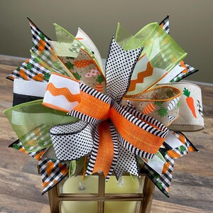 May include: A decorative Easter bow with various ribbons. The bow features orange, white, black, and green ribbons with carrot and polka dot patterns. The bow is placed on top of a wooden candle holder.