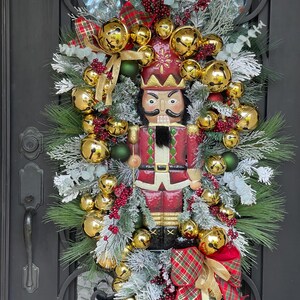 May include: A festive Christmas wreath featuring a red and gold nutcracker figure. The wreath is adorned with gold bells, red berries, green ornaments, and plaid bows. The greenery is accented with faux snow, creating a wintery holiday display.