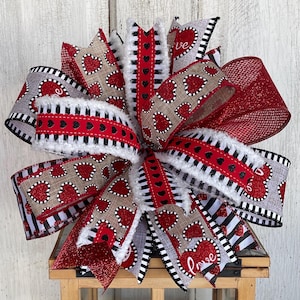 May include: A decorative bow featuring multiple ribbons in red, white, black, and gray. The ribbons are adorned with heart patterns, glitter, and the word "love". The bow has a fluffy, textured appearance, perfect for Valentine's Day or other romantic occasions.