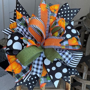 May include: A large, decorative bow made of black and white polka dot, black and white buffalo plaid, and orange and white pumpkin print ribbon. The bow is tied with a green burlap ribbon and has a rustic, farmhouse style.