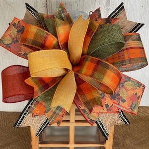 May include: A decorative fall-themed bow featuring a variety of ribbons in autumn colors. The bow includes ribbons with plaid, leaf, and solid patterns in shades of orange, green, and gold. The bow is attached to a wooden lantern.