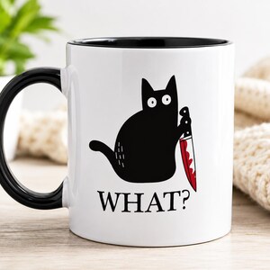 May include: A white ceramic mug with a black handle and interior. The front features a graphic of a black cat with wide eyes holding a knife with a red blade. Below the cat, the word WHAT? is printed in bold black capital letters.