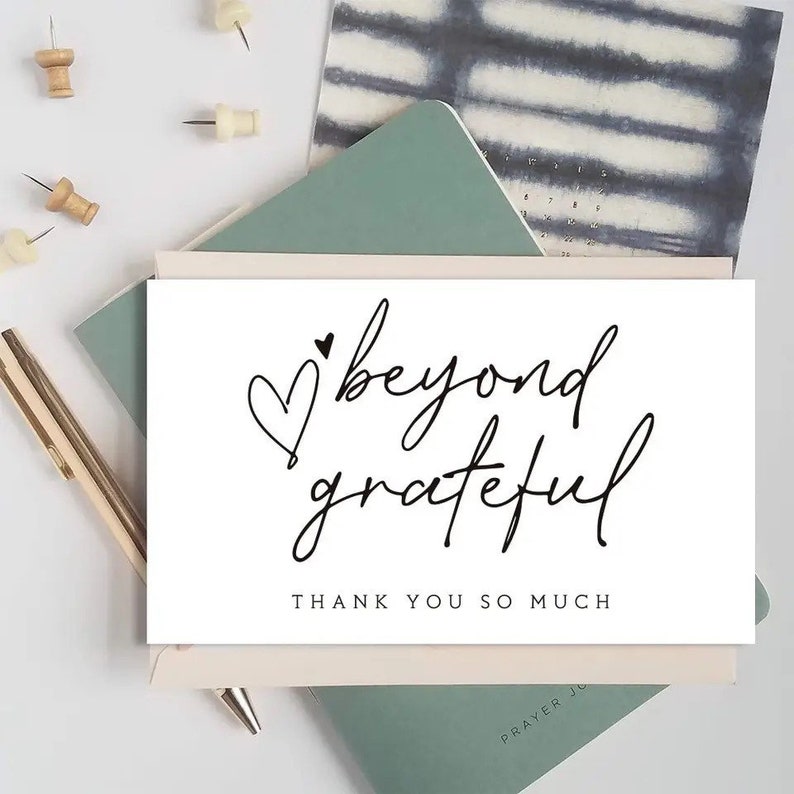 May include: A white card with the words "beyond grateful" in elegant black script, accompanied by a heart symbol. The phrase "THANK YOU SO MUCH" is printed below. The card is placed on a desk with a pen, push pins, and a notebook.