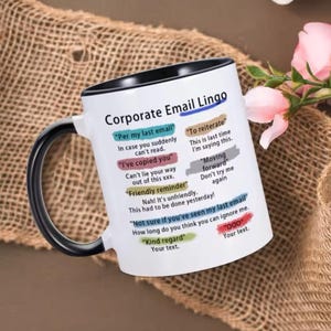May include: White ceramic coffee mug with a black handle. The mug has a black rim and features a humorous guide to corporate email lingo with phrases like "Per my last email", "I've copied you", "Friendly reminder", "To reiterate", "Moving forward", "Not sure if you've seen my last email", and "Kind regard".