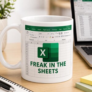 May include: White ceramic mug with an Excel spreadsheet design, the Microsoft Excel logo, and the text "FREAK IN THE SHEETS". The mug is on a wooden desk, with a laptop and notepad visible in the background.