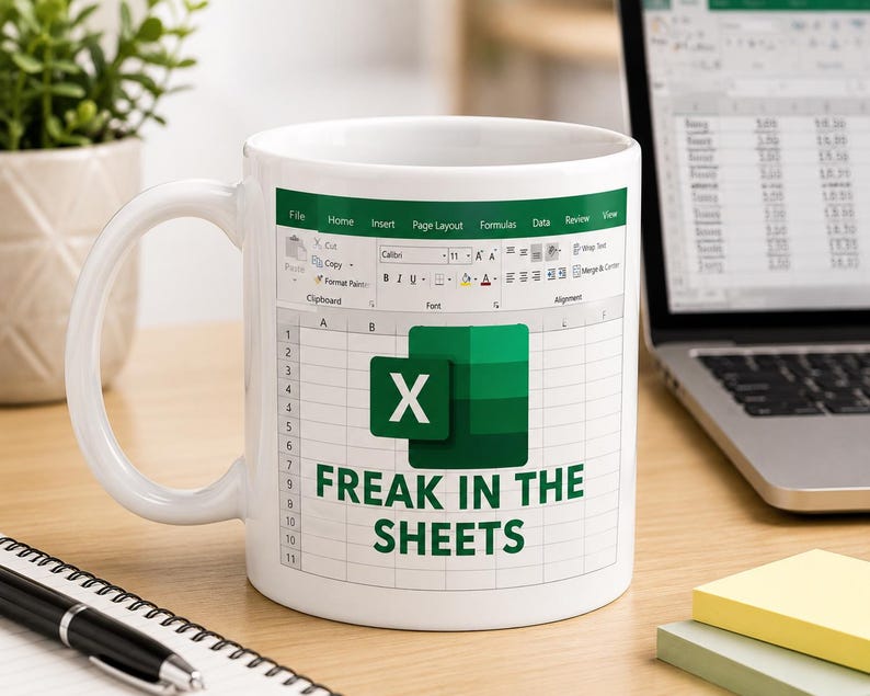 May include: White ceramic mug with an Excel spreadsheet design, the Microsoft Excel logo, and the text "FREAK IN THE SHEETS". The mug is on a wooden desk, with a laptop and notepad visible in the background.