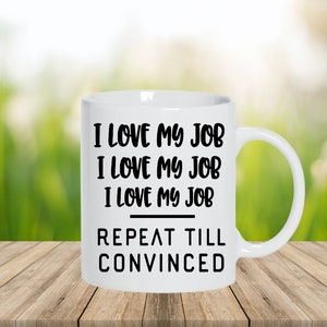May include: White ceramic mug with the text "I LOVE MY JOB" repeated three times, followed by "REPEAT TILL CONVINCED" in black lettering. The mug has a curved handle and sits on a wooden surface against a blurred green background.