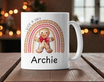May include: White ceramic mug featuring a gingerbread man design with a rainbow and the text "I BELIEVE IN SANTA" and "Archie". The mug has a curved handle and is set on a wooden surface. Festive holiday theme.
